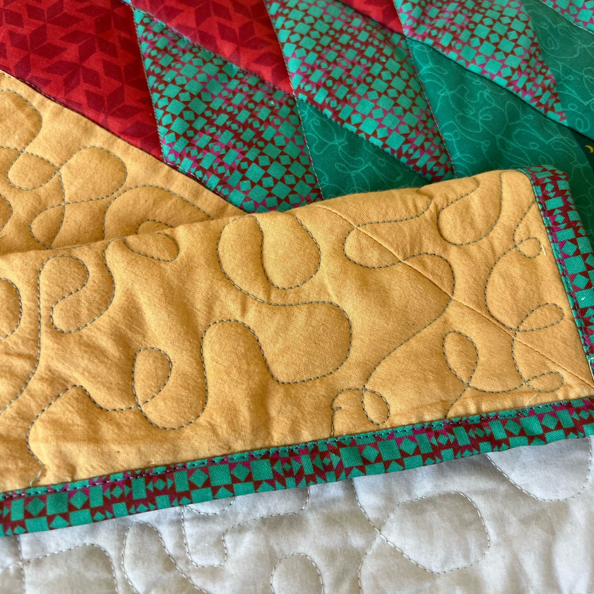Close-up of a quilted textile with a mustard yellow section featuring abstract stitched outlines, bordered by red and teal patterned panels, and a white quilted section visible at the bottom.