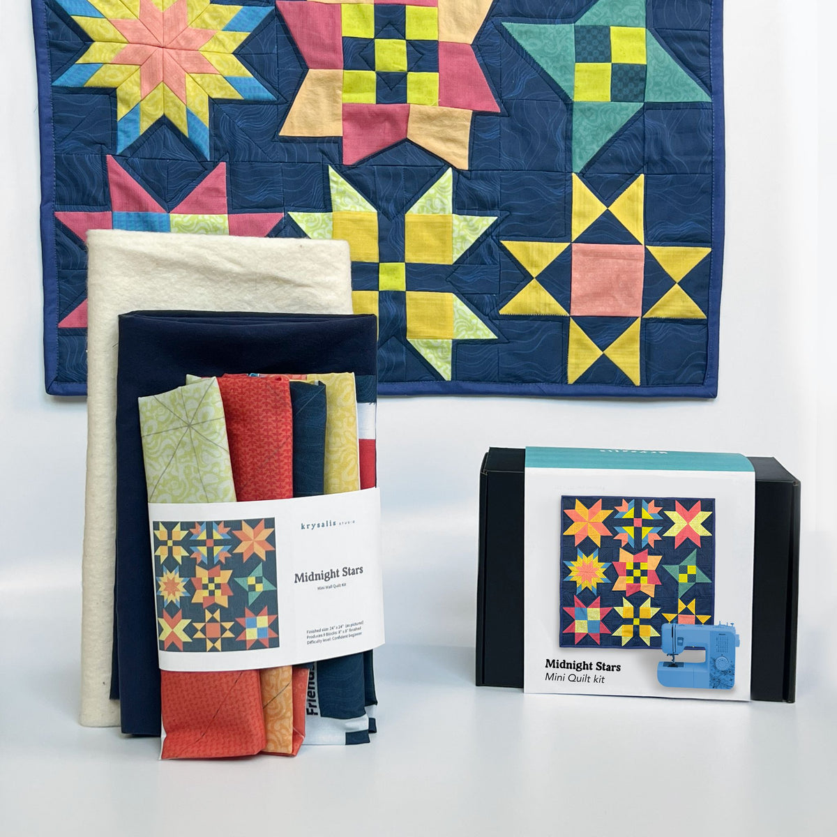 A quilting kit display featuring a folded bundle of fabric with a 'Midnight Stars' pattern label, a boxed 'Midnight Stars Mini Quilt Kit', and a large quilt showcasing the 'Midnight Stars' pattern in navy, yellow, pink, and green.