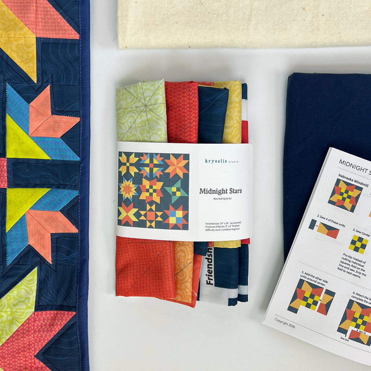 A printed panel fabric bundle wrapped in a paper band labeled 'krysalis Midnight Stars Quilt kit' is arranged on a white surface alongside a finished quilt, a folded navy blue fabric piece, and a printed ilustrated instruction sheet.