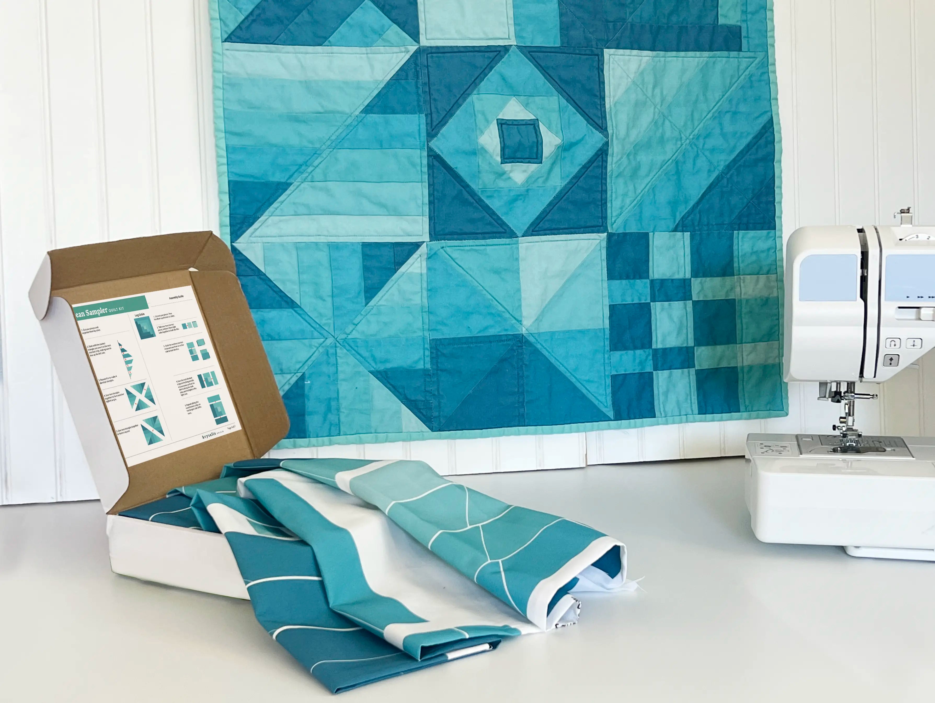 Quilt with geometric pattern, fabric roll, and sewing machine on a white surface.