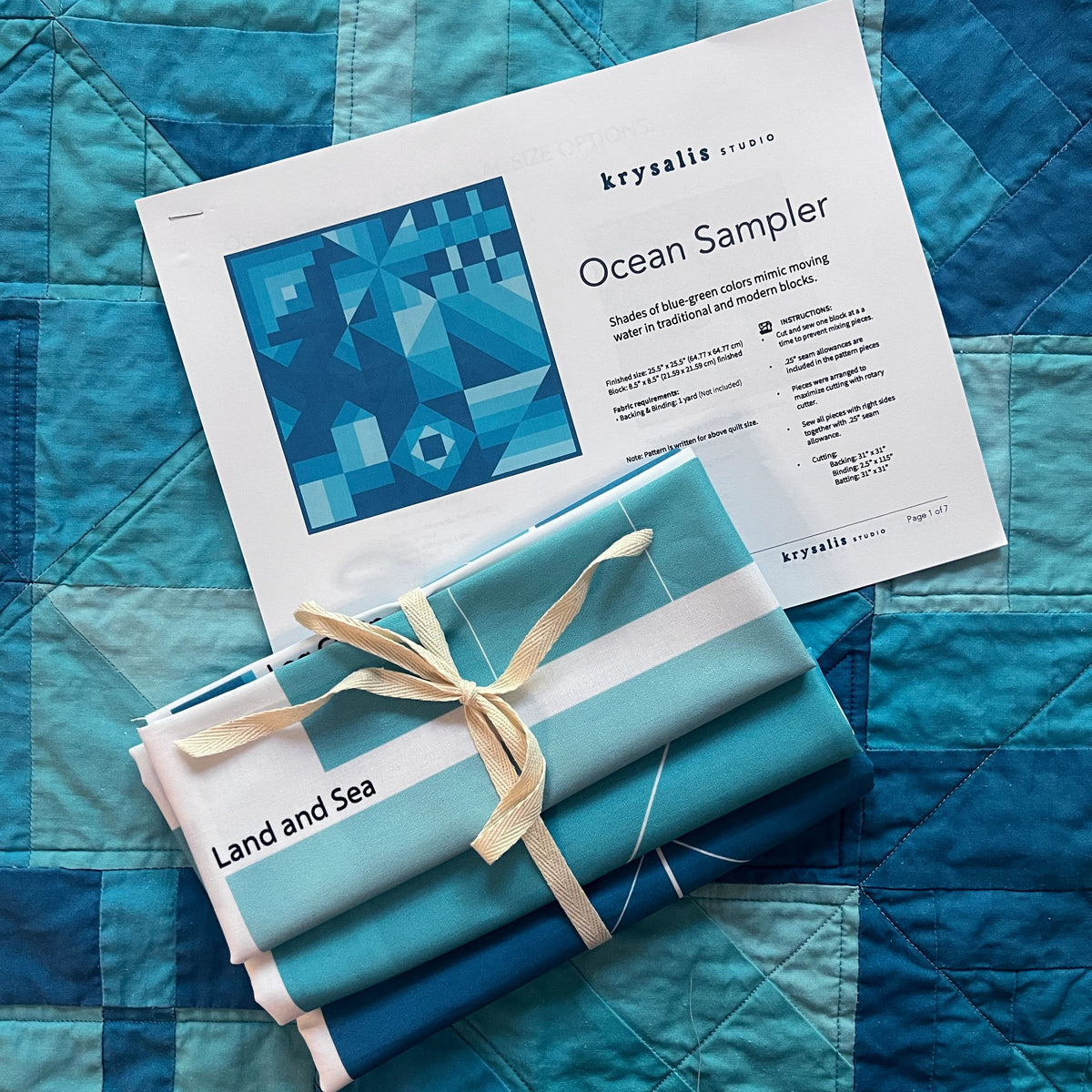 A styled flat-lay of a quilting fabric bundle wrapped in a cream ribbon, placed on top of a printed pattern sheet titled 'Ocean Sampler' by krysalis STUDIO. The fabric bundle contains shades of blue, teal, and white, labeled 'Land and Sea'.