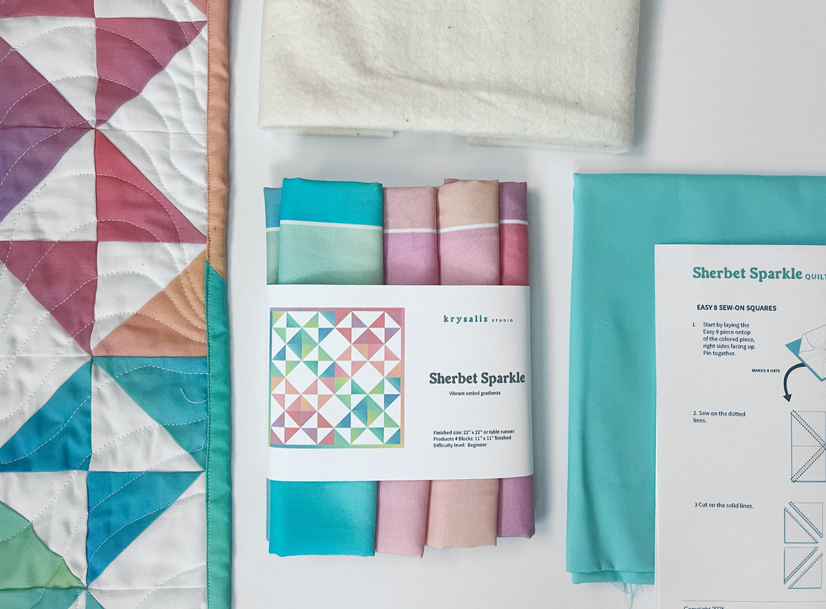 A flat-lay product presentation of Krysalis's Sherbet Sparkle quilt kit, showing a finished quilt with teal, pink, and purple geometric patterns, a folded white fabric piece, the packaged fabric bundle with a paper label, and a printed instruction sheet on a white background.